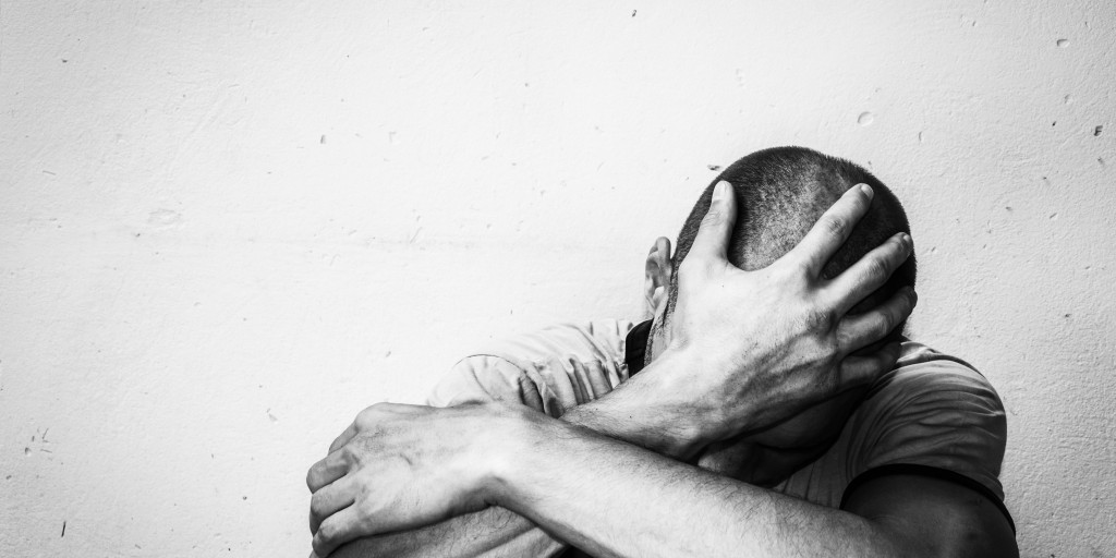 greyscale photo of distressed man covering head and slumping against wall in shame due to relapse