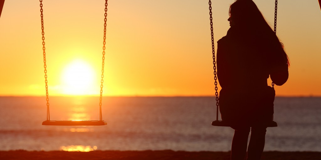 woman sitting alone on a swingset looking out at sunset on the ocean